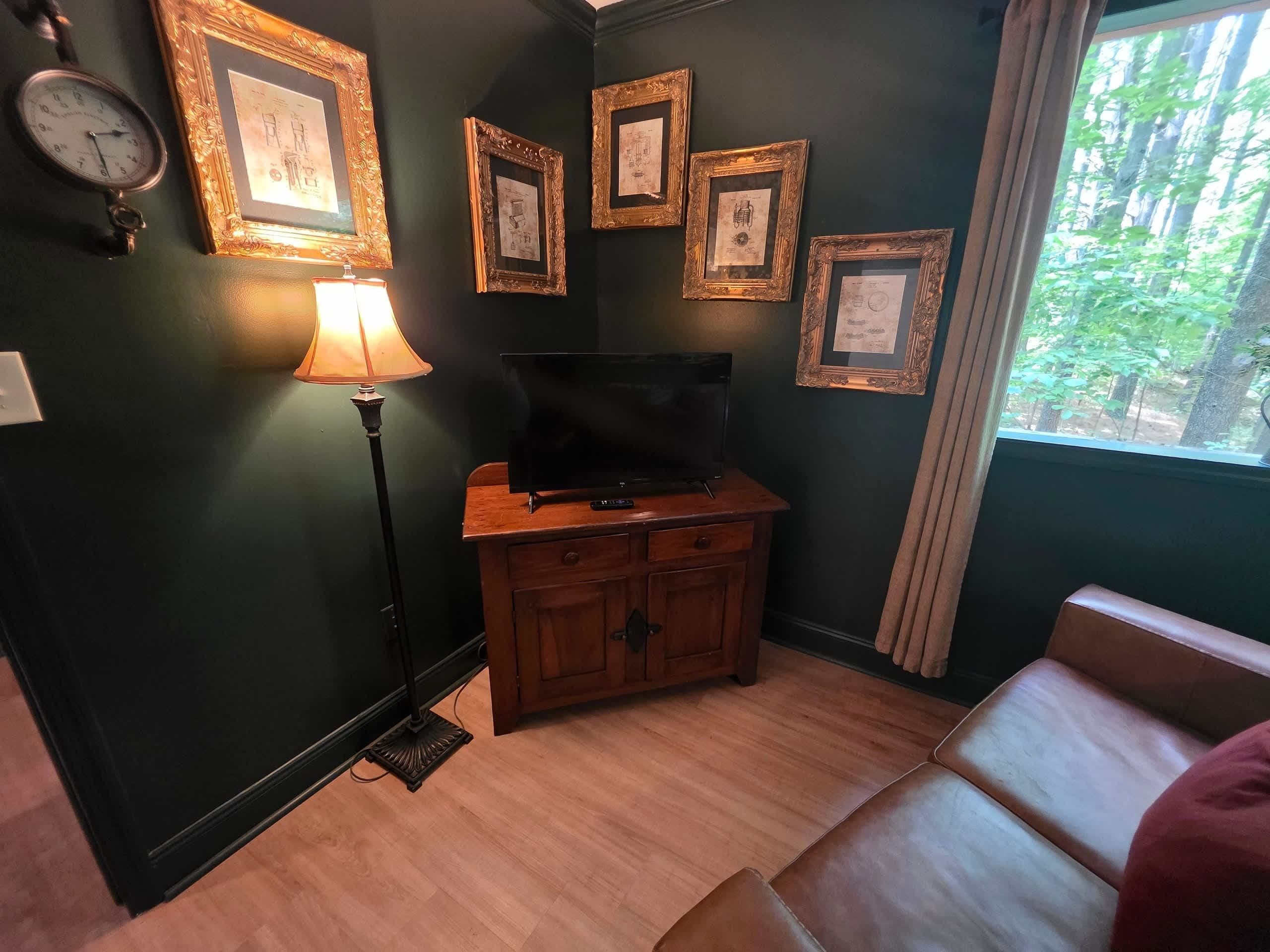 Een kamer met een televisie op een houten kast, ingelijste foto's aan de muur, een vloerlamp en een raam met uitzicht op bomen.