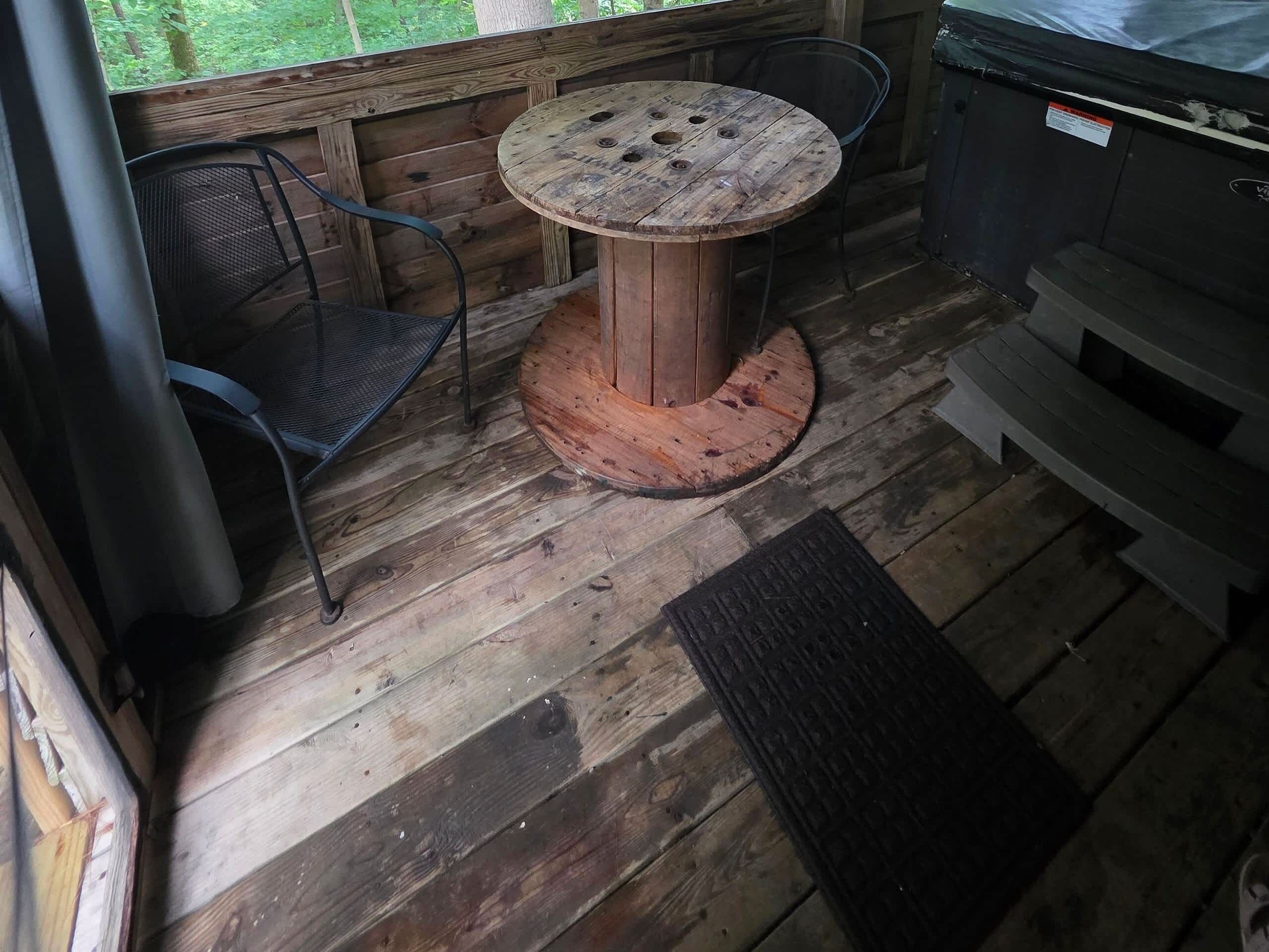 Een houten terras met een ronde tafel en stoelen, een deurmat en een vuilnisbak.