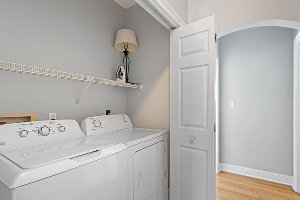 Un espace buanderie équipé d’un lave-linge et d’un sèche-linge à chargement frontal, d’une étagère avec une lampe et d’une porte donnant sur une autre pièce.