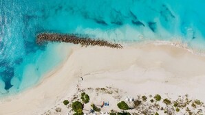 Vista aerea di una spiaggia con un molo roccioso, acque cristalline turchesi e una zona resort con ombrelloni e lettini.