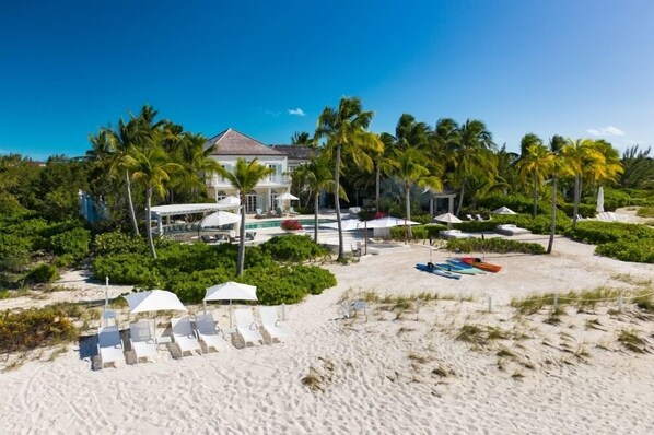 A beachfront property with a large house, palm trees, and beach chairs.