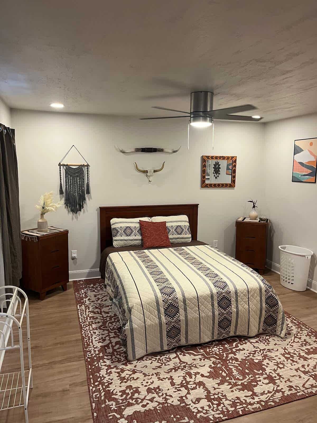 A bedroom with a bed, a ceiling fan, a wall-mounted macrame, a wall-mounted skull, and a framed abstract painting.