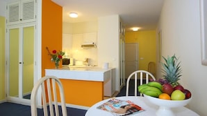 A compact kitchen with white cabinets, a white countertop, and a small island. A dining table with a magazine and a fruit bowl is in the foreground. - Caniço Bay Apartments Studio 1 (Caniço)