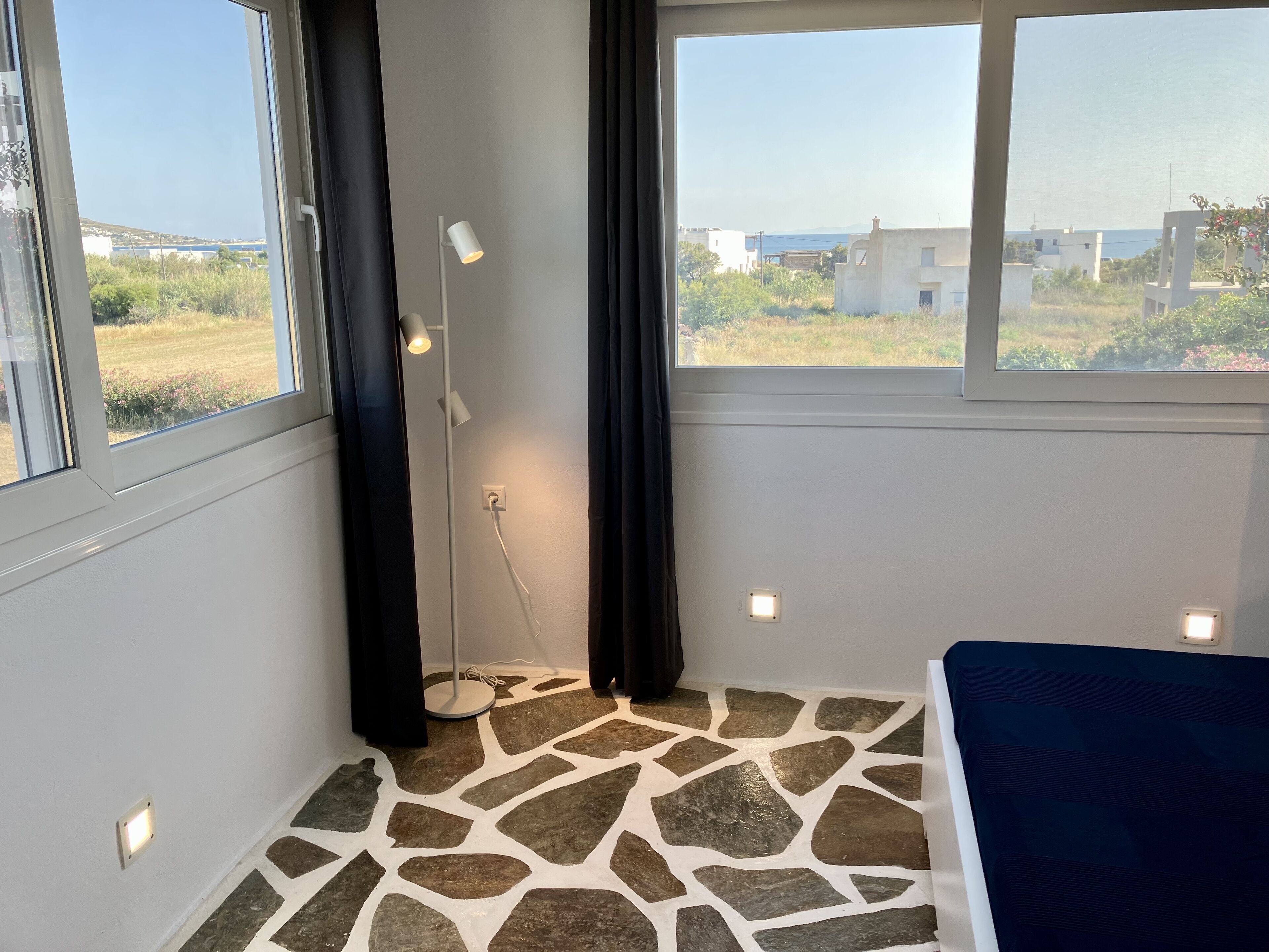 A room with a stone-patterned floor, a bed, a window with a view of buildings and greenery, and a standing lamp.