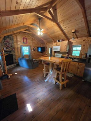 A cabin with a wooden interior, a fireplace, a kitchen area, and a dining table. - Buckeye Hut at Avenwood Cabins (new straitsville)