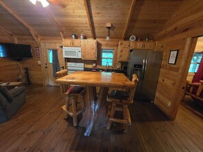 L’intérieur d’une cabane en bois comprend une table à manger centrale, des chaises en bois, une cuisine avec un réfrigérateur et un téléviseur fixé au mur.
