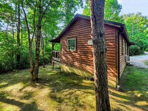 A wooden cabin with a porch, surrounded by trees and a grassy area.