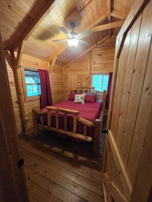 Ein Schlafzimmer einer Holzhütte mit einem Bett, einem Deckenventilator und zwei mit Vorhängen versehenen Fenstern.
