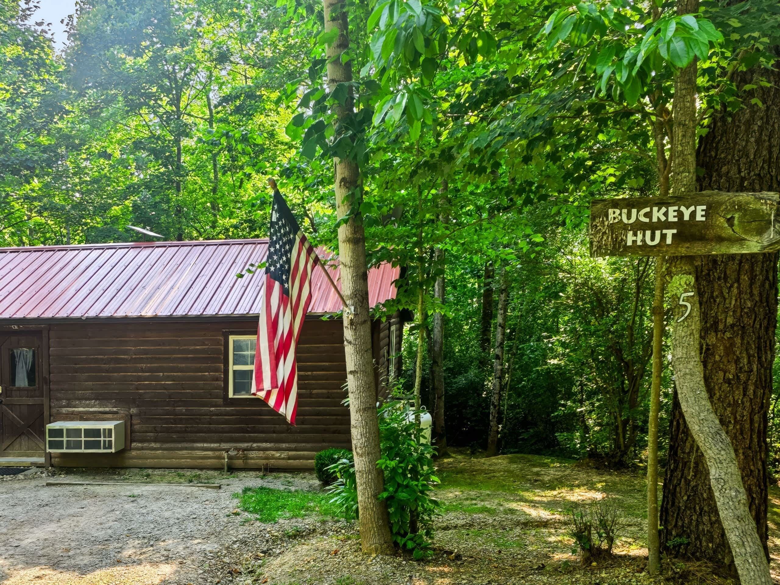 Eine rustikale Holzhütte mit amerikanischer Flagge, umgeben von dichtem Wald, und einem Schild mit der Aufschrift 'BUCKEYE HUT 5'.