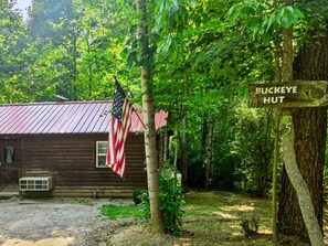 Une cabane rustique, ornée d’un drapeau américain, entourée d’une forêt dense, et sur laquelle est apposée une pancarte indiquant « BUCKEYE HUT 5 ».