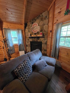 A cozy log cabin interior with a stone fireplace, a couch, and Ohio State memorabilia. - Buckeye Hut at Avenwood Cabins (new straitsville)