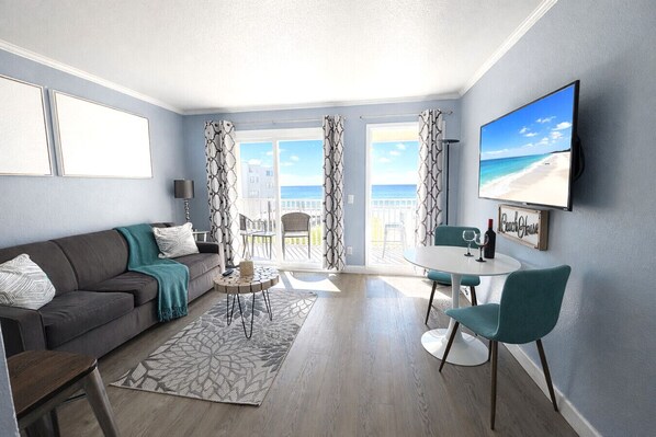 Un salón moderno con sofá, mesa de centro y sillas, un balcón con vistas al mar y un televisor de pantalla plana en la pared.