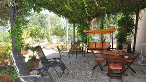 Een terras buiten met een ronde tafel en stoelen, omringd door potplanten en een pergola die schaduw biedt.