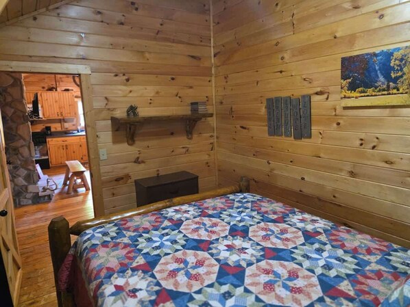 A wooden cabin interior with a bed, a wooden bench, a small table, and a stone wall leading to a kitchen area.