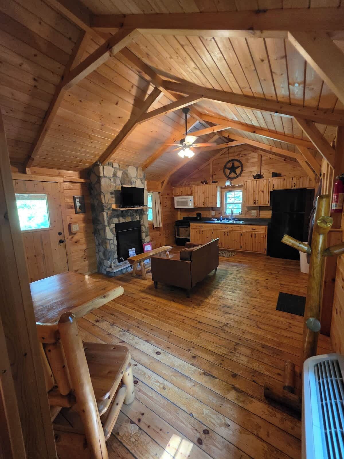 Interno di una baita di legno con camino in pietra, arredi in legno, angolo cottura e ventilatore a soffitto.
