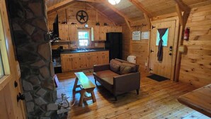Ein gemütliches Blockhaus-Interieur mit Kamin aus Stein, Holzdielenboden, einer Couch und einer Küchenzeile.