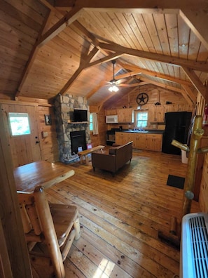 Un acogedor interior de cabaña con chimenea de piedra, muebles de madera, una cocina con microondas y un ventilador de techo.