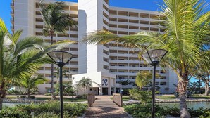 A multi-story hotel with a central entrance, surrounded by palm trees and a pathway leading to the entrance. - Riviera Beach Club 502 - 2B/2B Dream Vacation Condo w/ Amazing Gulf Views! (Fort Myers Beach)