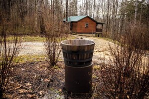 Une cabane rustique située dans une zone boisée, avec un baril en métal au premier plan.