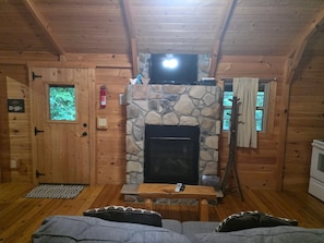A cabin interior with a stone fireplace, wooden walls, a television mounted above it, a small wooden table, and a sofa.