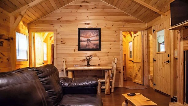 A wooden cabin interior with a leather sofa, a dining table, and a fireplace. - Whippoorwill at Avenwood Cabins (new straitsville)