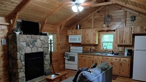 A cozy cabin interior with a stone fireplace, wooden walls, a kitchenette, and a sofa. - Whippoorwill at Avenwood Cabins (new straitsville)
