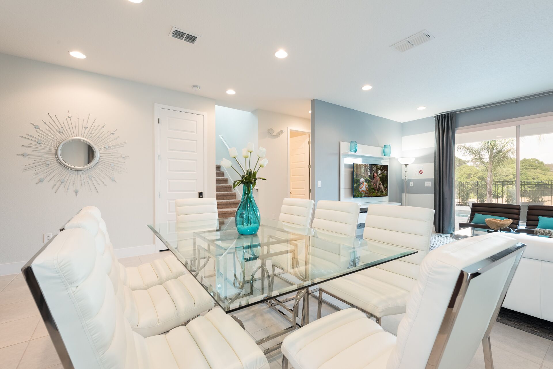 A modern dining area with a glass table and white chairs, a teal vase with flowers, and a view of the outdoors.