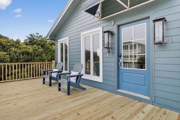 Une maison bleue avec une terrasse, deux chaises Adirondack et une porte avec une fenêtre.