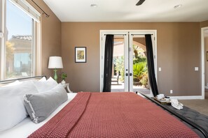 A hotel room with a bed, a window, a door leading to a balcony, and a small table with a lamp and a picture frame.