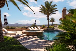 A pool area with lounge chairs and umbrellas, surrounded by palm trees and a view of mountains.