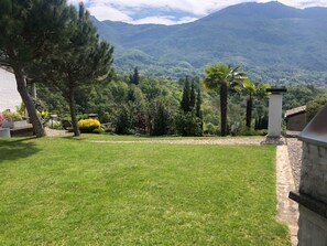 Une pelouse bien entretenue, avec un chemin de pierres menant à un bâtiment, le tout entouré de divers arbres et dominé par un paysage montagneux.