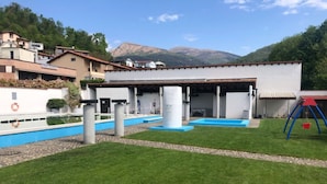 A swimming pool area with a covered deck, a swing set, and a grassy lawn. - Sureggio Village (Capriasca)