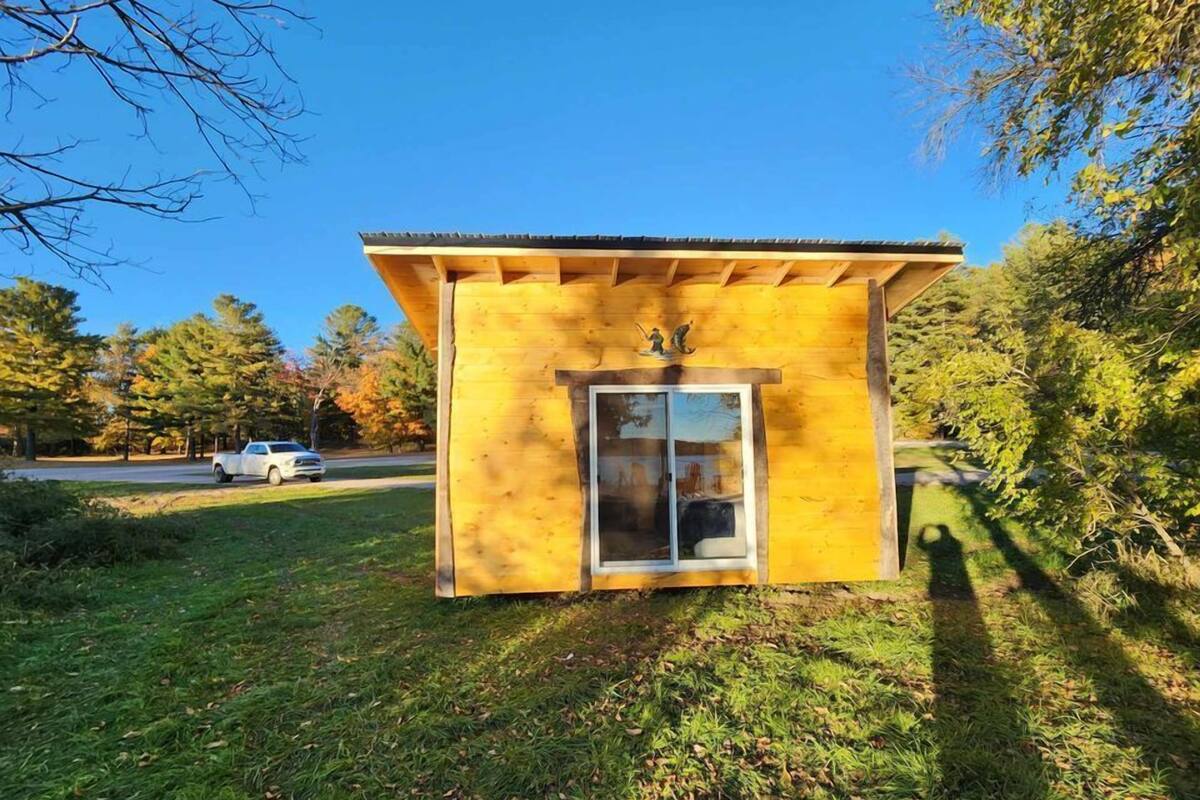 Una pequeña cabaña de madera con puerta de cristal y techo metálico, rodeada de árboles y un coche estacionado.