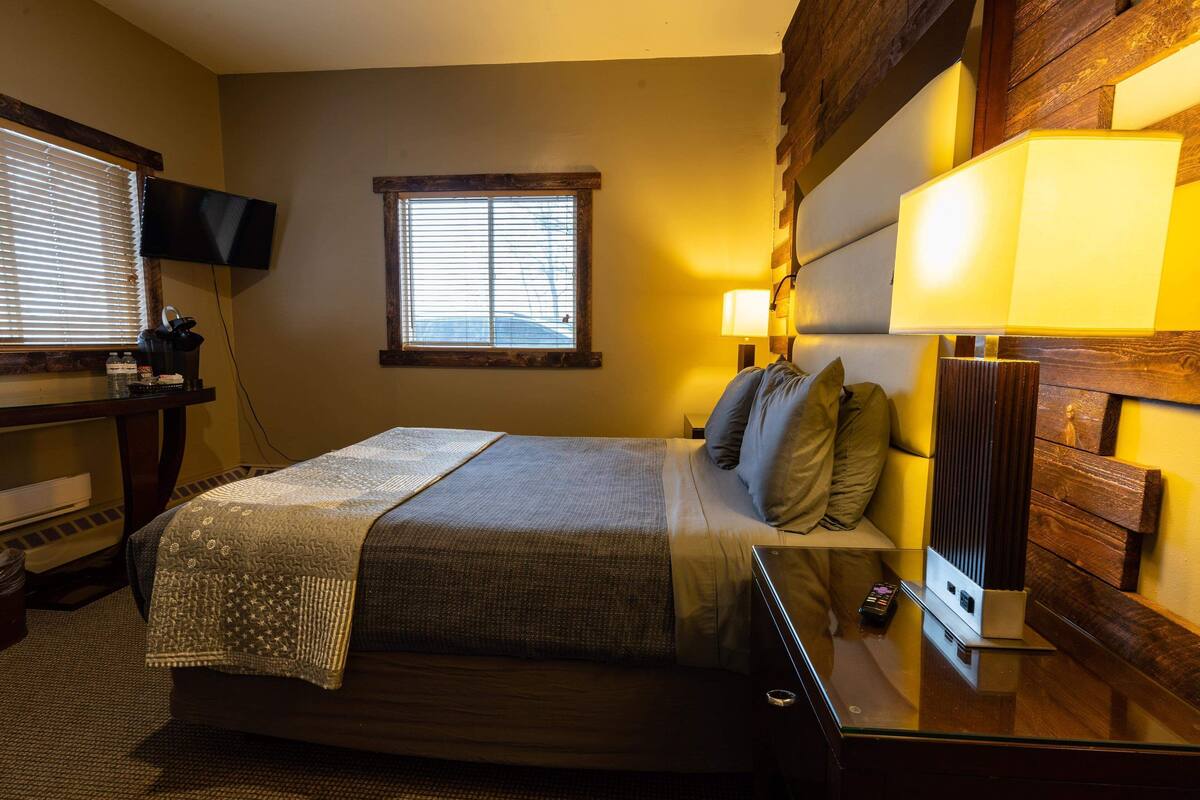 Habitación de hotel con cama, mesita de noche, lámpara, televisor y ventana con persianas.
