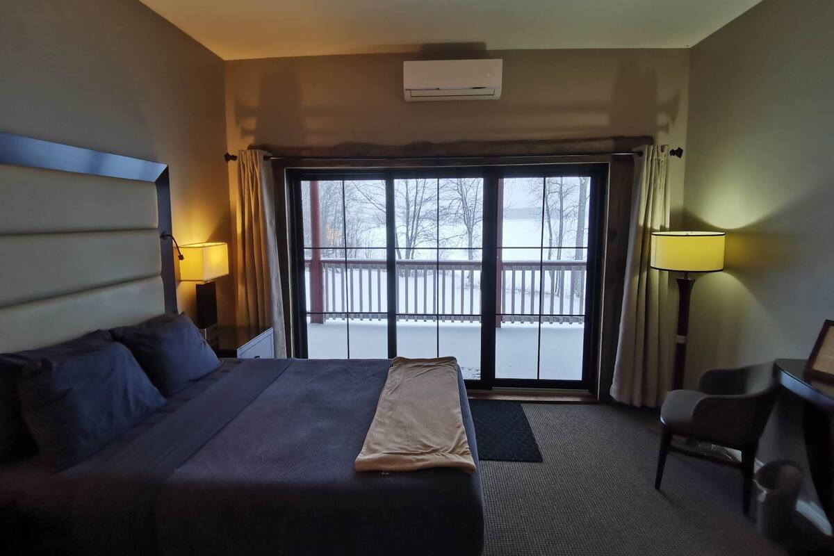 Habitación de hotel con cama, escritorio, una silla, un ventanal con vistas a una terraza nevada y dos lámparas.