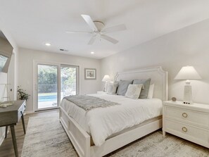Une chambre à coucher avec un lit, des tables de chevet, un ventilateur de plafond et une vue sur l’extérieur, visible à travers une porte vitrée.