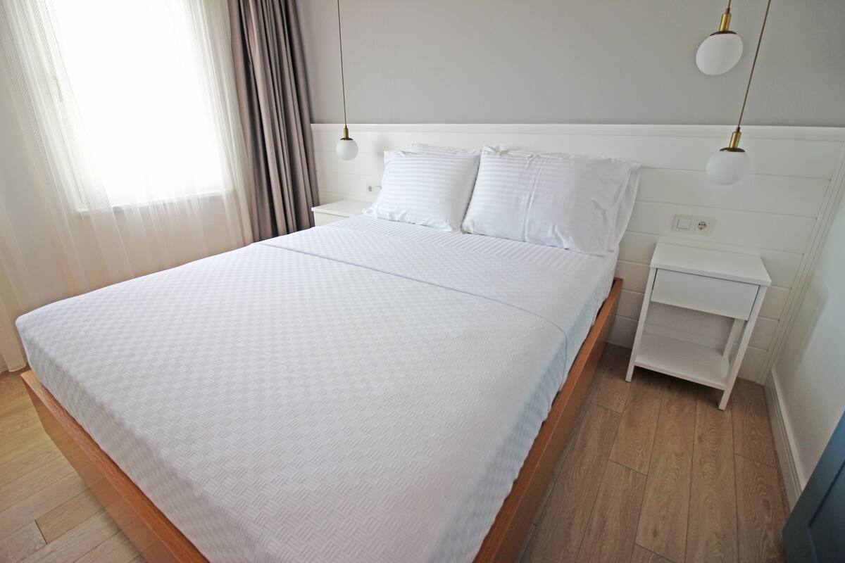 A neatly made bed with white linens, a bedside table, and a nightstand with a lamp.