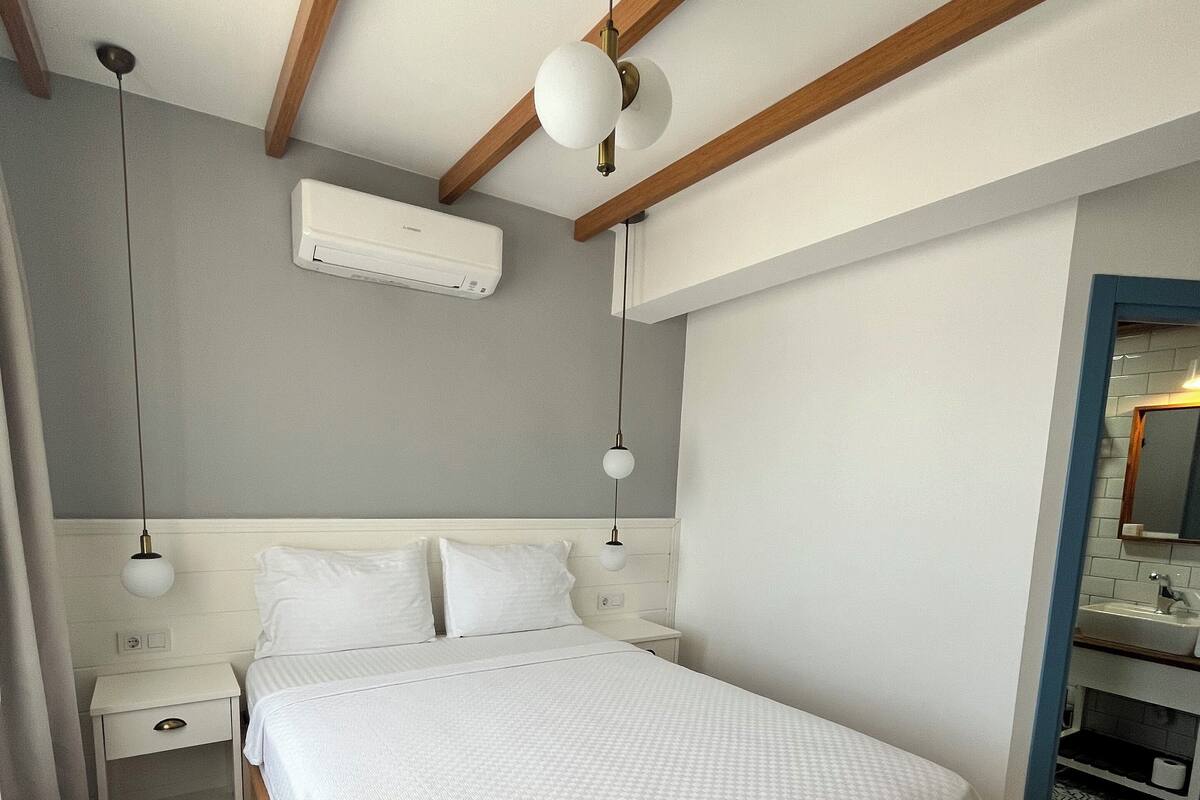 A bedroom with a large bed, wooden beams, a white air conditioner, bedside tables, and a mirror in the corner.
