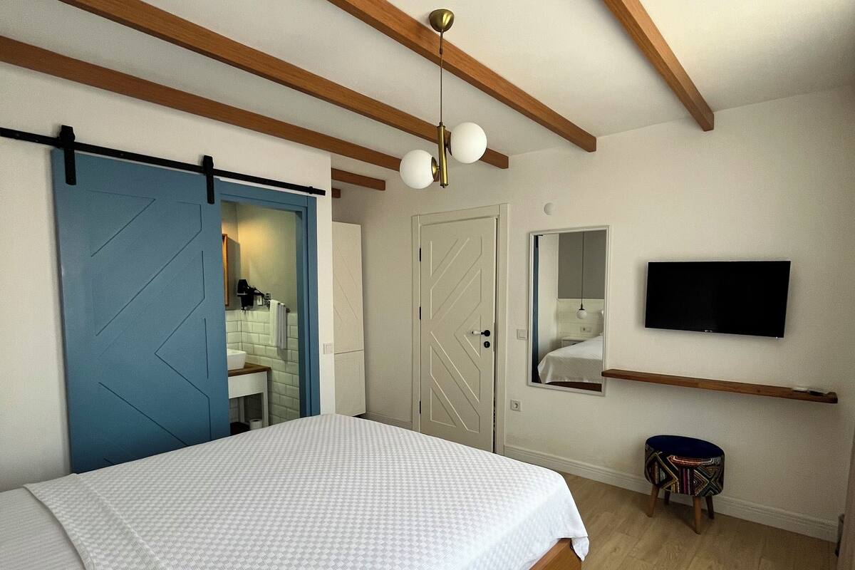 A bedroom with a bed, wooden beams, a ceiling light, a TV, and a small stool.