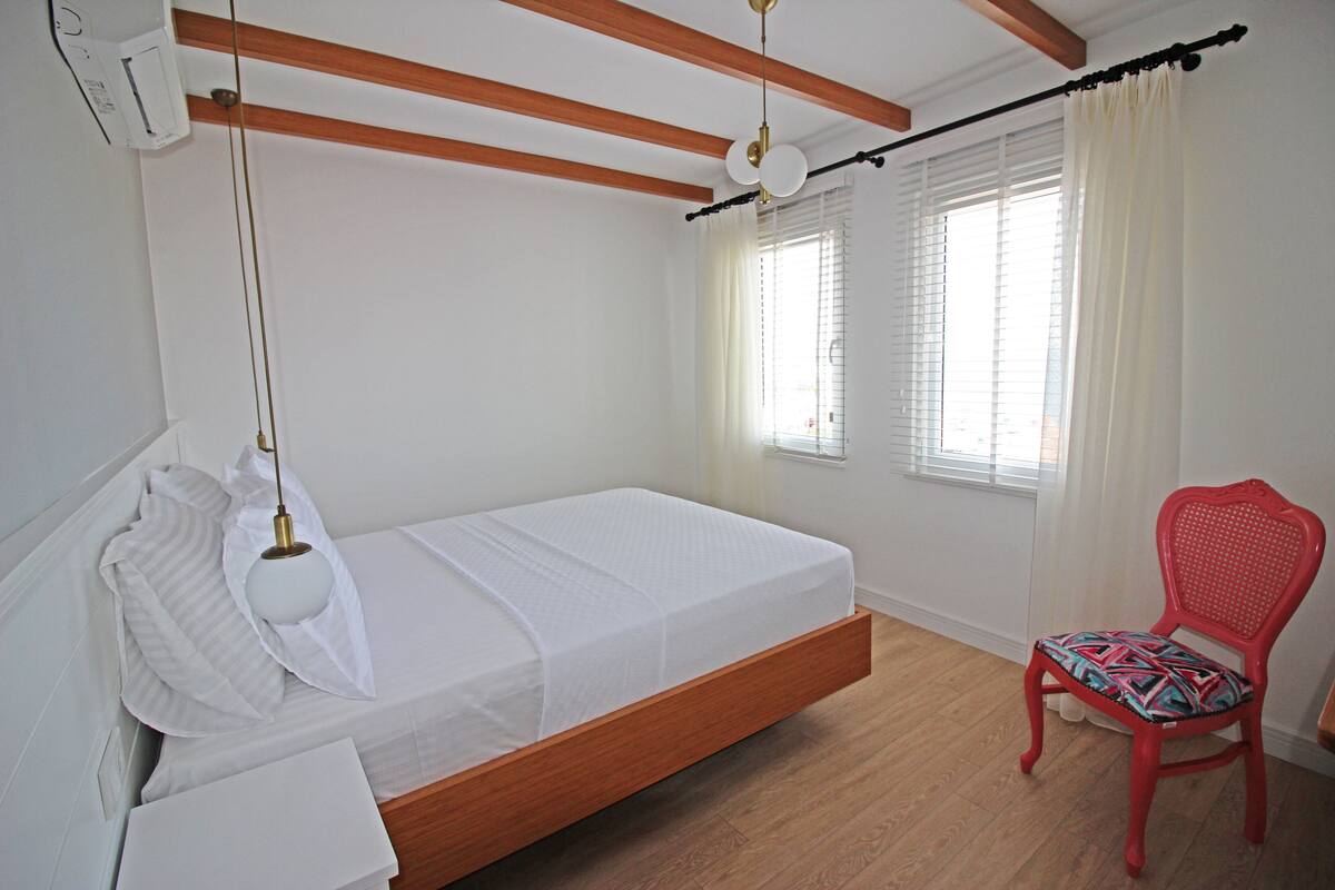 A bedroom with a wooden bed frame, white bedding, a red chair with patterned cushion, and a window with white curtains.