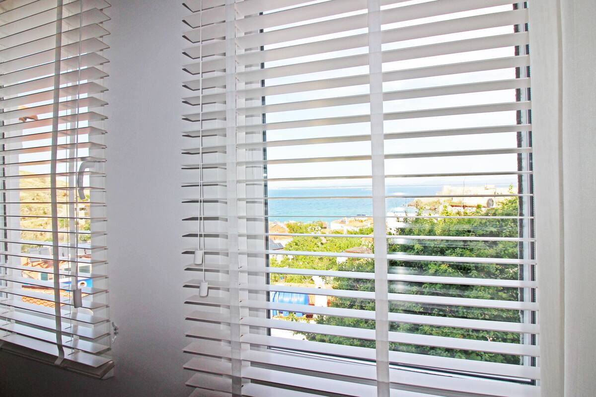 A window with horizontal blinds, a view of the sea, and greenery in the background.