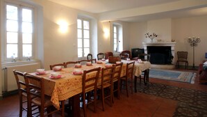 Une salle à manger avec une grande table dressée pour six personnes, entourée de chaises en bois. On y trouve une cheminée avec un manteau de cheminée, ainsi qu’une fenêtre donnant sur l’extérieur.