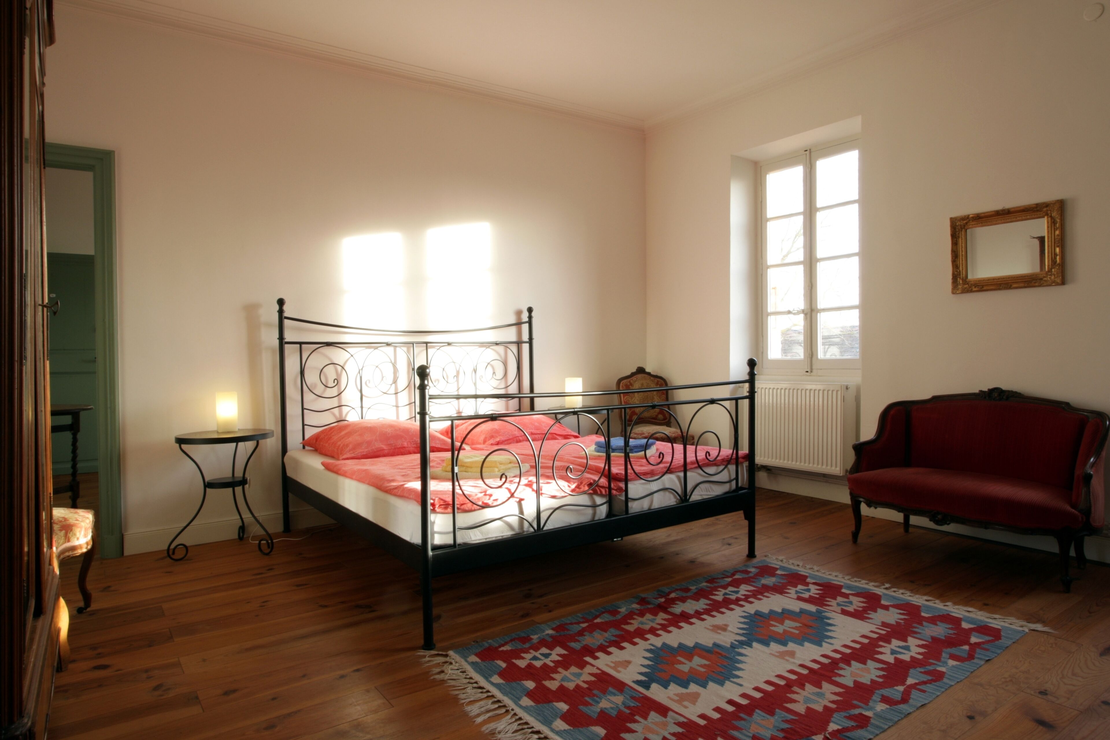 Une chambre à coucher avec un lit en métal, un canapé rouge, un tapis à motifs et une fenêtre laissant entrer la lumière naturelle.
