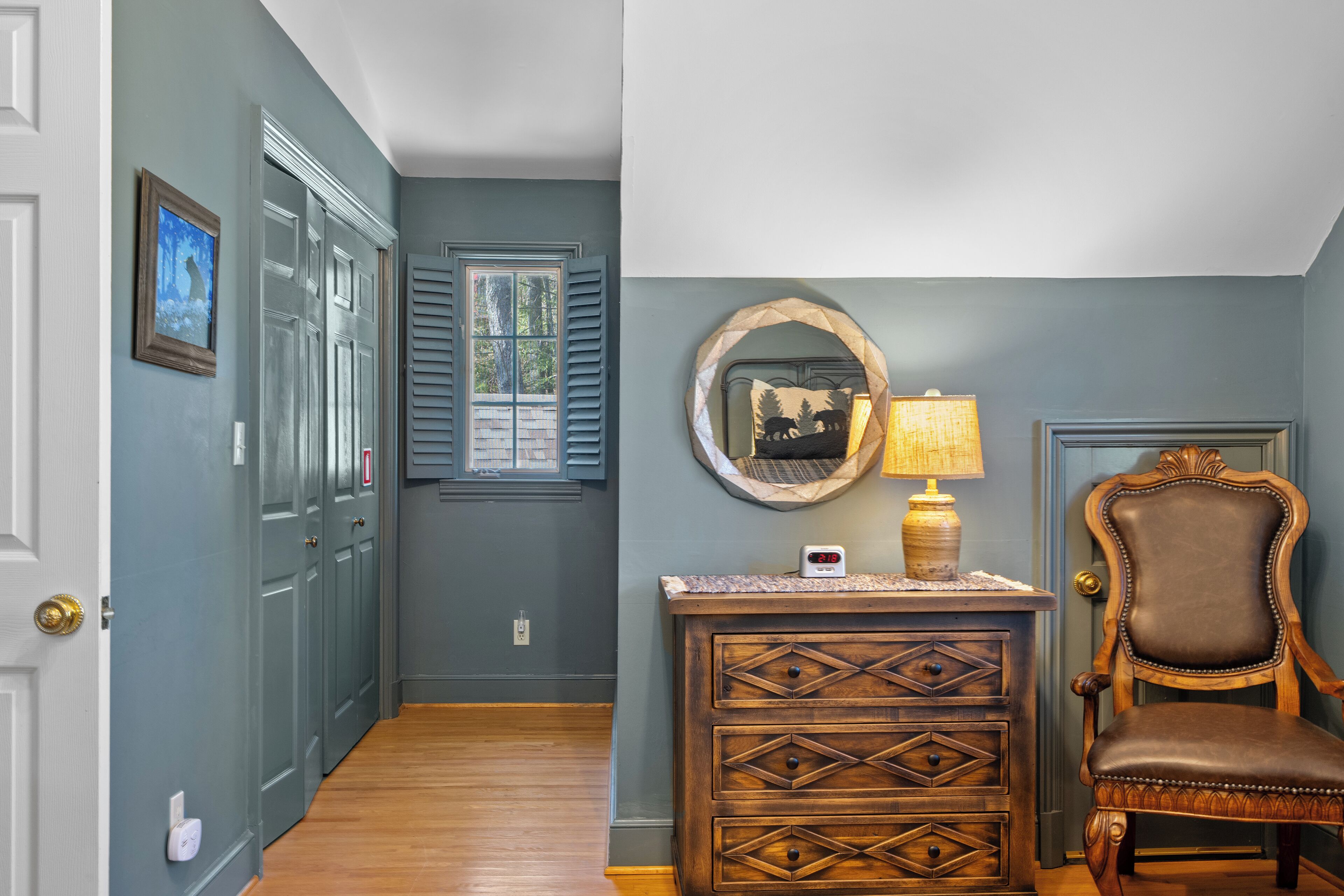 A hallway with a wooden floor, a door, a window with shutters, a wall-mounted mirror, a lamp, and a chair.