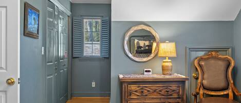 A hallway with a wooden floor, a door, a window with shutters, a wall-mounted mirror, a lamp, and a chair.
