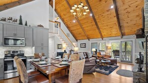 A spacious living area with a high ceiling, wooden beams, a dining table set for four, and a cozy seating arrangement. - Smoky Mountain Cabin with Stunning National Park Views (Gatlinburg)