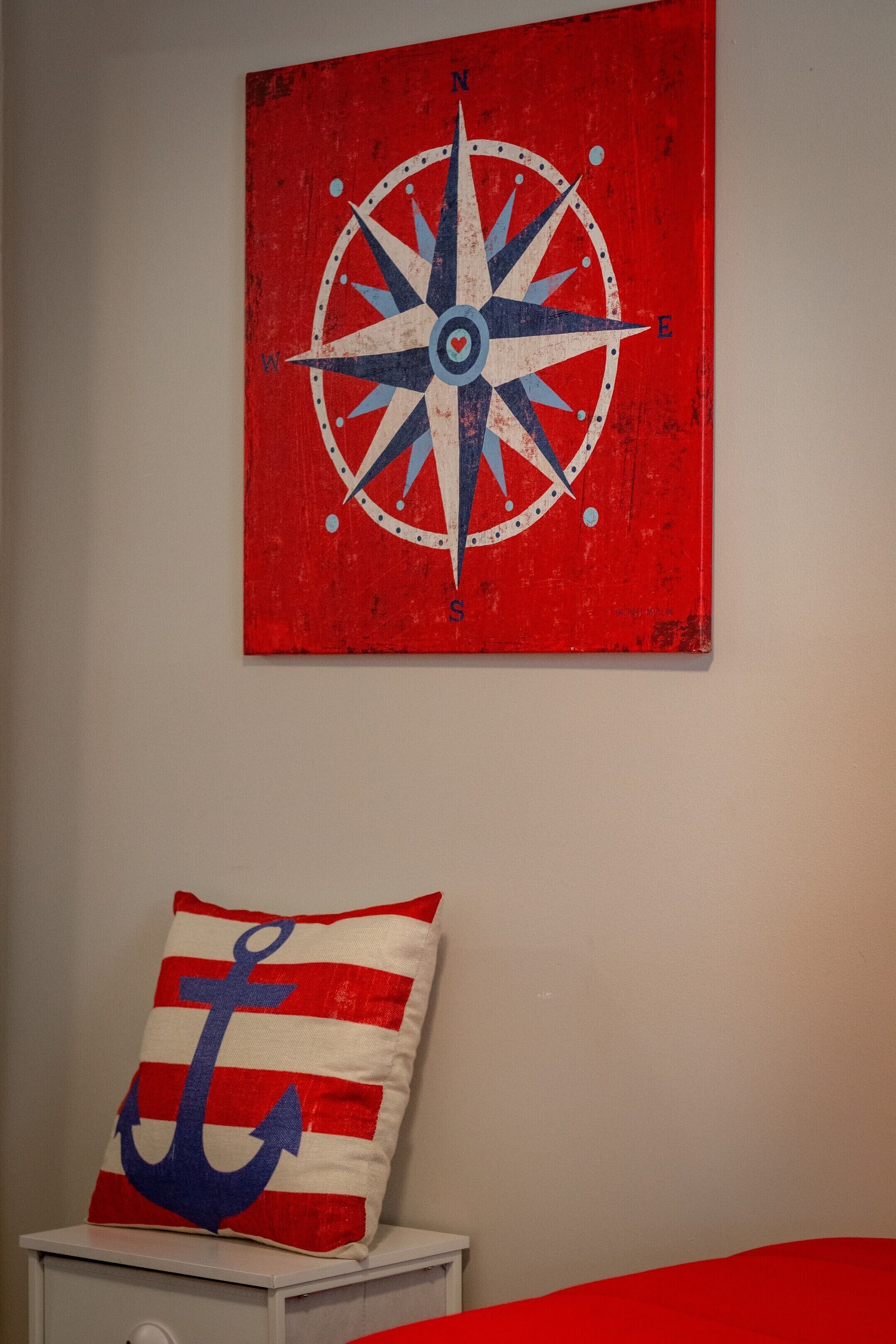 A room with a red wall, a white nightstand, a red and white pillow with an anchor design, and a red and white striped pillow.