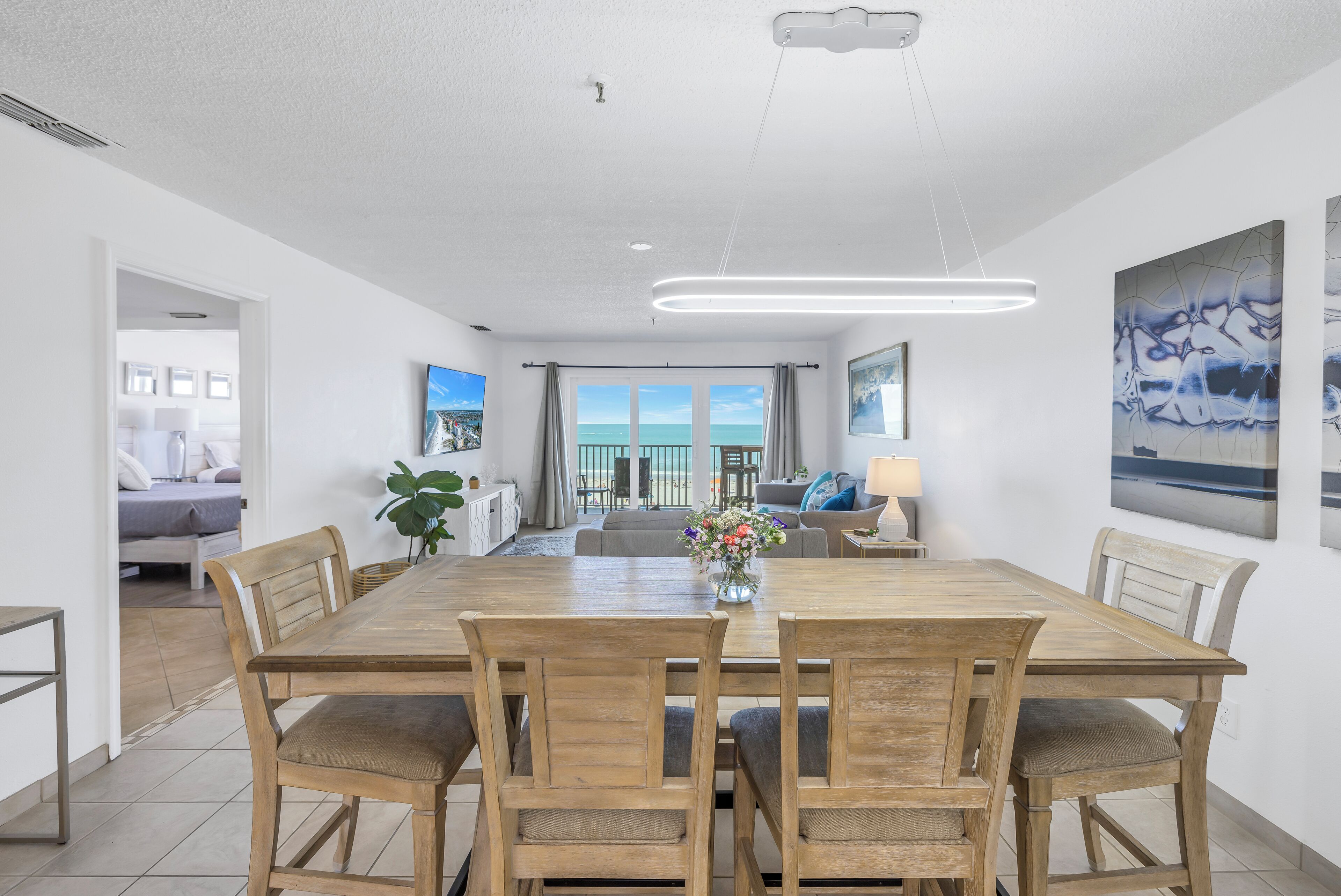 A spacious dining area with a long wooden table and chairs, a vase of flowers, and a view of the ocean through large windows.