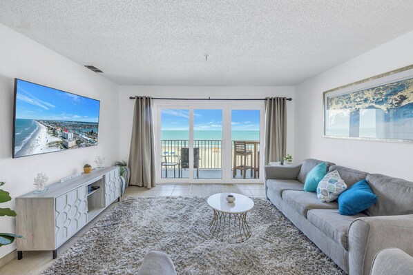A modern living room with a large TV, a grey sofa, and a view of the beach through the sliding glass doors.