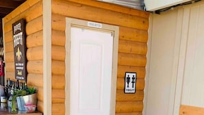 A wooden cabin with a white door, a sign for a toilet, a small table with plants, and a large pumpkin outside.
